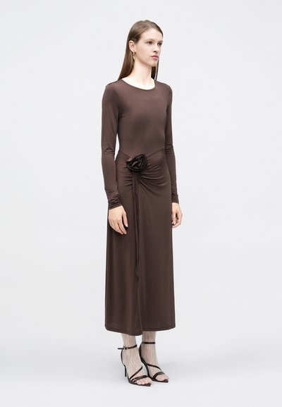 Γυναίκα που στέκεται φορώντας καφέ midi φόρεμα με μακριά μανίκια, με σούρα στη μέση και floral λεπτομέρεια, φορώντας δαντελένιες κάλτσες και μαύρες ψηλοτάκουνες σαγιονάρες με λουράκια.