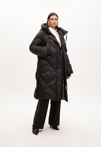 Černý, huňatý dlouhý kabát s kapucí, s diamantovým prošíváním, obsahující zipové kapsy a akcenty leopardího vzoru uvnitř.