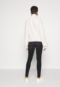 Jersey de punto crema con cuello alto y mangas abullonadas, combinado con jeans ajustados negros y zapatillas blancas con detalles amarillos.