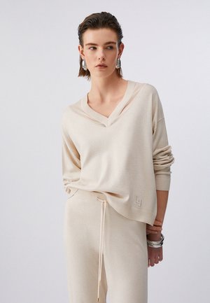 Donna che indossa un maglione a V color crema e pantaloni abbinati con coulisse, abbinati a orecchini e braccialetti d'argento, su sfondo neutro.