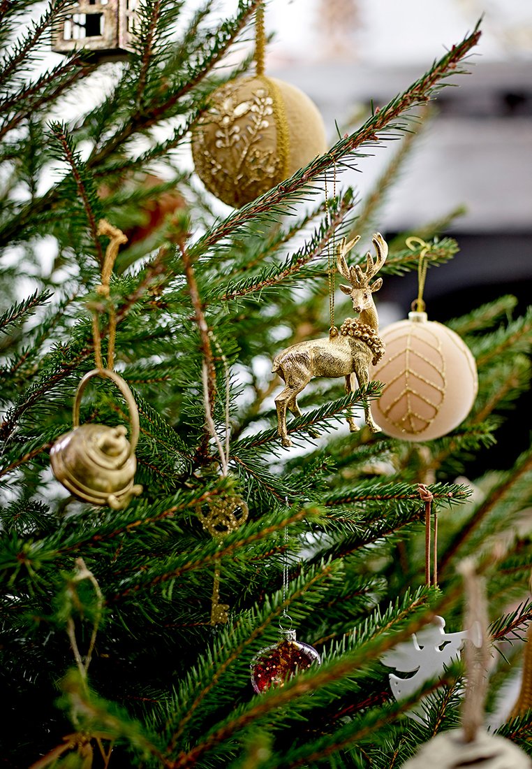 Weihnachtsbaum geschmückt mit goldenen Ornamenten, einem goldenen Rentier, dekorativen Glocken und weißen Schneeflocken, umgeben von grünen Nadeln.