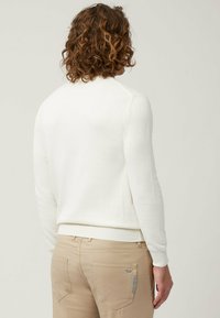 Felpa bianca a maniche lunghe con collo rotondo, polsini e orlo a costine. Indossata con pantaloni beige chiaro con tasche posteriori.