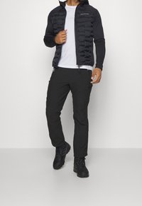 Veste matelassée noire avec fermeture éclair à l'avant, associée à un pantalon stretch noir et des chaussures de randonnée. Texture lisse, légère et design fonctionnel.