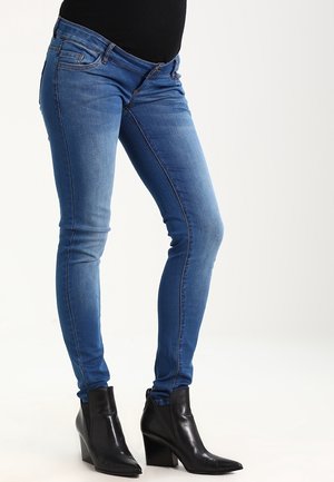 Personne portant un jean skinny bleu et des bottines noires à talons, debout de profil sur un fond uni.
