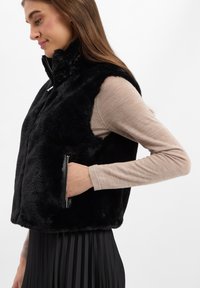 Czarny kamizelka z sztucznego futra z wysokim kołnierzem, zapięciem na zamek i kieszeniami bocznymi. Noszona na jasnobeżową bluzkę z długim rękawem i plisowaną czarną spódnicę.