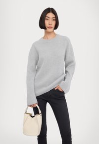 Pulover ribbed gri deschis cu mâneci lungi, purtat cu blugi negri. Modelul ține o geantă mică albă tip bucket cu un breteau.