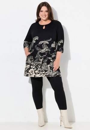 Schwarze Tunika mit Paisleymuster und beigen Blumenakzenten, Dreiviertelärmel, Schlüsselloch-Ausschnitt, kombiniert mit schwarzen Leggings und cremefarbenen Stiefeletten.