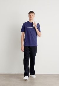 Tricou bleu marin cu "BARE SUN INTERNATIONAL" imprimat în alb, asortat cu pantaloni negri și adidași albi, ținând un prosop drapat pe umăr.