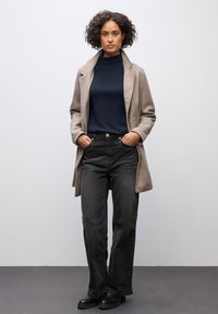 Beige Wollmantel über einem navyfarbenen Rollkragenpullover, kombiniert mit schwarzen Jeans mit hoher Taille. Das Model steht mit den Händen in den Taschen und trägt schwarze Stiefeletten.