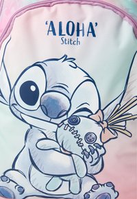 Stitch-Charakter, der eine mit Knopfaugen und einer Schleife verzierte Puppe umarmt, unter dem Text "'ALOHA' Stitch" auf einem pastellrosa Hintergrund.