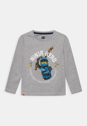 Grå langærmet skjorte med blå ninjafigur, der holder et våben, to gule stjernedolk, og tekstene "Ninja Hero" og "Master Your Ninja Journey".