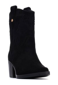 Botín de ante negro con tacón cuadrado, que presenta detalles de costura, punta redondeada y tiradores en el caña para un fácil calzado.