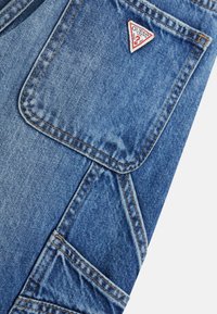 Pantalons cargo en denim bleu moyen avec une finition texturée, dotés d'un logo de marque triangulaire rouge, de multiples poches et de coutures contrastées.