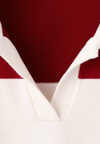 Primo piano di un colletto bianco a maglia che si apre su uno sfondo di tessuto a maglia rosso con una striscia orizzontale rossa visibile sotto il colletto.