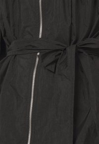 Vêtement d'extérieur noir à finition texturée, équipé d'une fermeture éclair frontale et d'une ceinture nouée à la taille. Le tissu semble légèrement froissé.