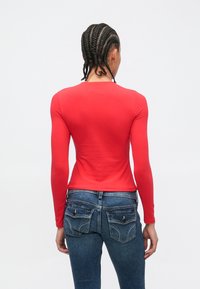 Person med flettet hår iført en rød, tettsittende genser med lange ermer og blå jeans, stående med ryggen til mot en enkel hvit bakgrunn.