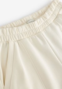 Cream-colored sweatpants with an elastic waistband, featuring side pleats and a soft fabric texture. Label visible inside waistband.