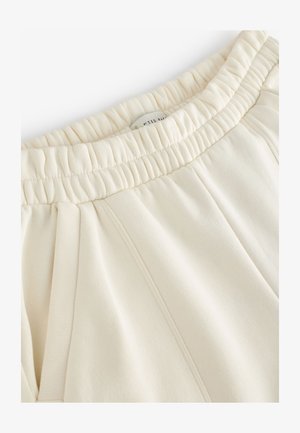 Kremo spalvos sportinės kelnės su elastinga juosmens juosta, šoninėmis klostėmis ir minkšta audinio tekstūra. Etiketė matoma juosmens viduje.