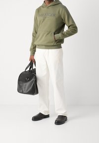 Felpa verde oliva con logo stampato, pantaloni bianchi a gamba larga, borsa in pelle nera con manici superiori e scarpe nere slip-on.