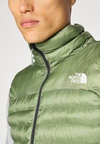 Groene puffer vest met een hoge kraag, gemaakt van glanzende stof. Beschikt over verticale stiksels en een wit logo op de linkerborst, met een zwarte rits.