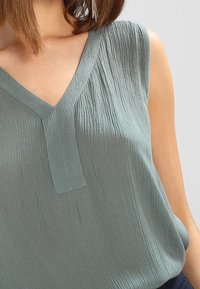Haut sans manches à col en V de couleur teal doux avec un tissu texturé et une légère brillance. Présente une couture centrale et une coupe décontractée.