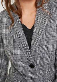 Marynarka w czarno-białą kraciastą kratę houndstooth, z kołnierzem z wykładanym i pojedynczym zapięciem na czarny guzik. Materiał o fakturze.