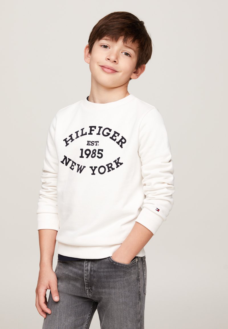 Felpa bianca con testo nero che legge "HILFIGER EST. 1985 NEW YORK," polsini a costine e un piccolo logo sull'orlo, abbinata a jeans grigi.