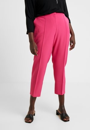 Person wearing bright pink cropped trousers, black long-sleeve top, and black open-toe block heel sandals standing on a white floor.