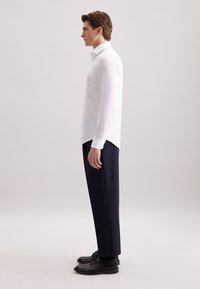 Jonge man in een wit overhemd met lange mouwen, donkere broek en zwarte nette schoenen, staande in profiel tegen een eenvoudige lichte achtergrond.