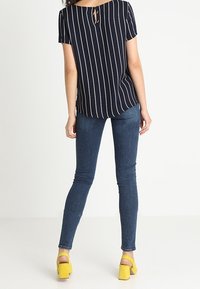 Frau, die ein schwarz-weiß gestreiftes Kurzarmoberteil, dunkelblaue skinny Jeans und gelbe Blockabsatzsandalen trägt, steht mit dem Rücken zur Kamera.