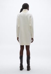 Fehér kötött pulóverruha magas gallérral, bordázott ujjakkal és egyenes sziluettel, térdig érő fekete bőr csizmával kombinálva.