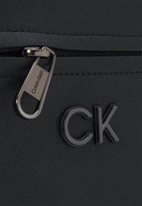 Borsa in finta pelle nera con chiusura a zip e una linguetta in metallo argentato con il logo "Calvin Klein" inciso. Il logo del marchio è ben visibile in nero.
