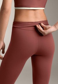 Primo piano di una persona che sistema la cintura di leggings color ruggine, indossando un top corto abbinato su uno sfondo neutro.