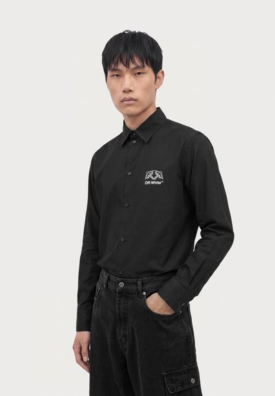 Homme portant une chemise noire à boutons avec le logo Off-White sur la poitrine et un pantalon cargo noir, debout avec une main dans la poche sur fond blanc.