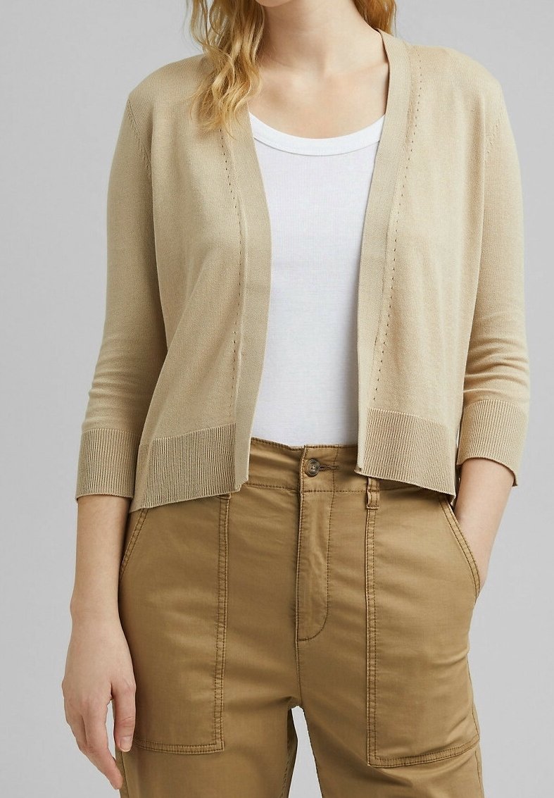 Femme portant un cardigan beige ouvert sur un haut blanc et un pantalon taille haute couleur beige, les mains dans les poches, sur fond gris uni.