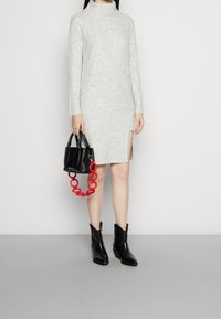 Abito a coste grigio chiaro con scollatura alta, spacco laterale, abbinato a una borsa nera con manico a catena rossa e stivaletti neri.