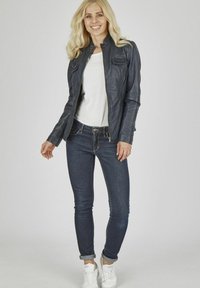 Dunkle Lederjacke mit Stehkragen, Reißverschluss und seitlichen Taschen, getragen über einem weißen T-Shirt und dunklen Jeans mit aufgeschlagenen Säumen. Weiße Turnschuhe.