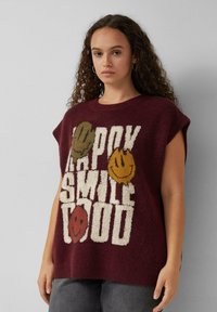 Frau mit lockigem Haar, die einen bordeauxfarbenen ärmellosen Pullover trägt, auf dem der Text "HAPPY SMILE GOOD" und drei bunte Smiley-Gesichter zu sehen sind.