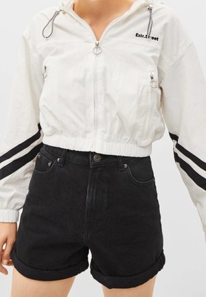 Veste blanche raccourcie avec des rayures noires, fermeture éclair et logo sur la poitrine, associée à des shorts en denim noirs taille haute retroussés aux ourlets.