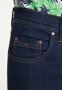 Ciemnoniebieskie dżinsy z klasycznym pięciokieszeniowym fasonem, kontrastującymi pomarańczowymi st stitching oraz metalowymi nitami na przednich kieszeniach.