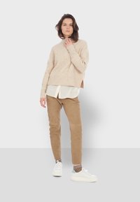 Beige geribde trui met zijsplitten, laag over een witte blouse. Tan suède broek en witte sneakers met blauwe accenten.