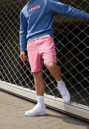 Personne portant un sweat bleu Levi’s, un short rose, des chaussettes blanches et des baskets blanches, debout avec une jambe levée près d’un portail en métal.