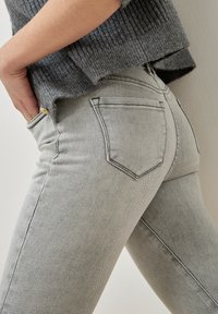 Primer plano de una persona que lleva unos jeans ajustados de color gris claro y un suéter de punto gris, con la mano descansando en la cadera.