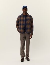 Brązowa i granatowa koszula w kratę na guziki, granatowy sweter pod spodem, szare spodnie i niebieska czapka z tekstem; czarne buty z grubą podeszwą.