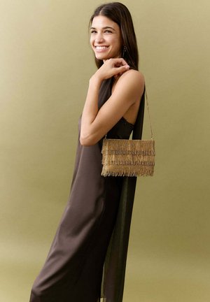 Femme souriante vêtue d'une robe marron foncé sans manches, tenant un sac à main à franges doré sur son épaule, sur un fond beige uni.