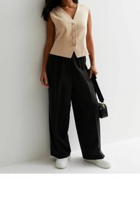 Beige ärmlös väst med V-ringning och fem knappar, ihopparad med löst sittande svarta byxor och vita sneakers. Liten svart handväska.