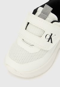 Sapato desportivo branco com parte superior em malha e tira de Velcro, apresentando aba cinzenta e logótipo "cK" preto na tira.