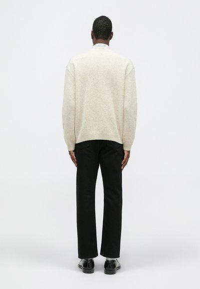 Bežs pārmērīgs džemperis ar ribotām apmales un piedurknēm, saskaņots ar melnajiem biksēm un melnajām kurpēm, redzams no mugurpuses uz balta fona.