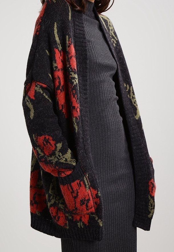 Cárdigan de punto con un fondo oscuro que presenta patrones florales en rojo y verde. Diseño de abertura en la parte delantera con mangas largas y un ajuste relajado.