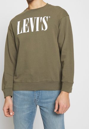 Mężczyzna w oliwkowej bluzie Levi's z dużym białym logo i jasnoniebieskich dżinsach, stojący na tle jasnego, jednolitego tła.